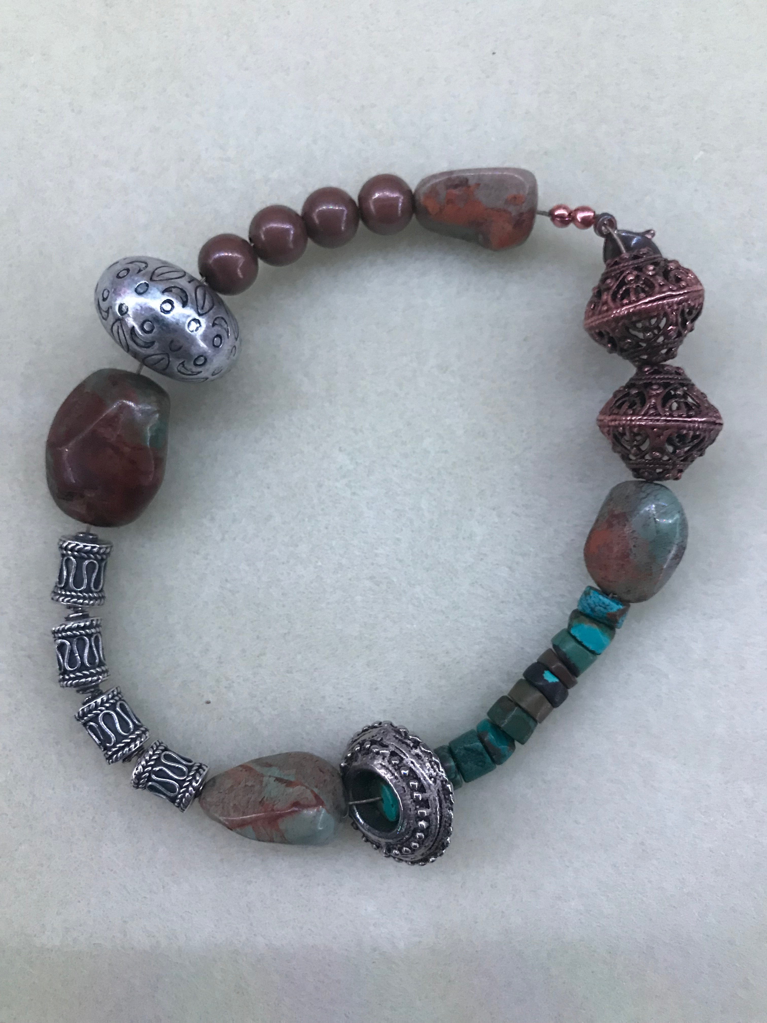 A stim loop made from brass ball bearing beads, turquoise heishi beads, copper basket beads, and irregularl shaped turqiouse beads with one flat side. Medium weight beading wire holds it all together. The loop rests on a white beading mat.